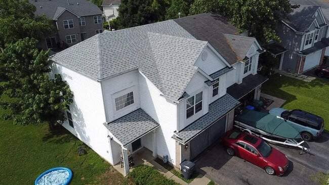 Aerial view of a two-story white suburban home with a newly installed light gray asphalt architectural shingle roof. The house features a multi-level design with a prominent front entrance and an attached garage. The photo includes a red sedan and a covered boat in the driveway, highlighting a complete, clean new roofing project.